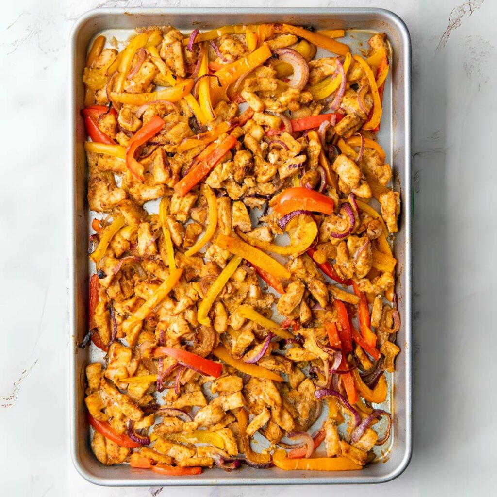 A sheet pan filled with cooked strips of chicken, yellow and red bell peppers, and red onions, all seasoned and evenly spread out.