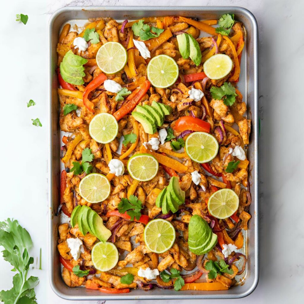 Sheet pan with cooked chicken, sliced bell peppers, red onions, topped with lime slices, avocado, cilantro, and dollops of sour cream.