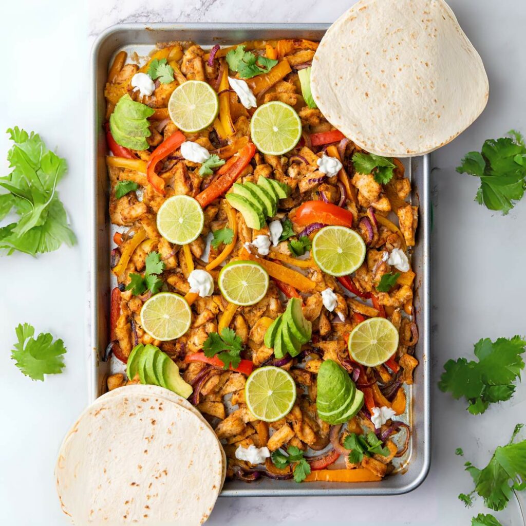 Sheet pan with cooked chicken, bell peppers, and onions, topped with lime slices, avocado, cilantro, and dollops of sour cream.