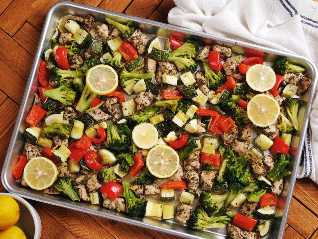 A sheet pan filled with baked chicken pieces, broccoli, zucchini, red bell pepper, and lemon slices on a wooden surface.