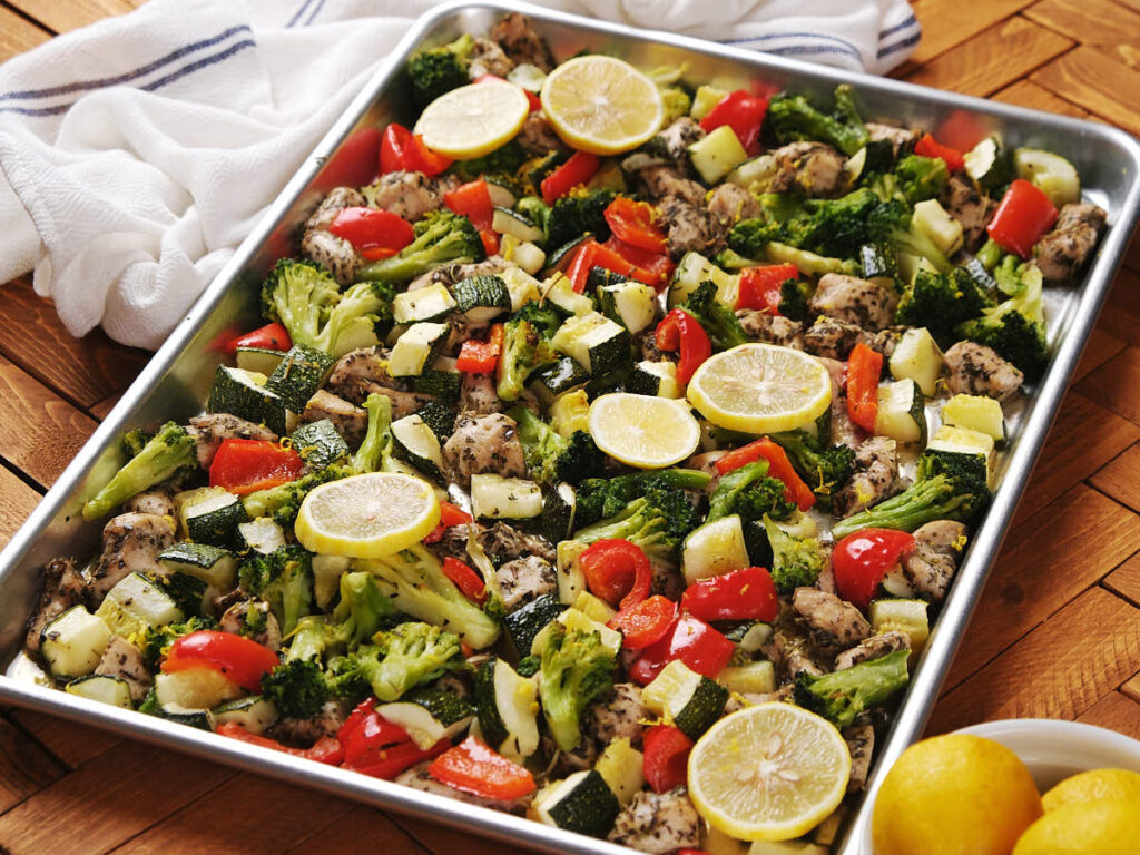 A sheet pan filled with baked chicken pieces, broccoli, red bell peppers, zucchini, and lemon slices on a wooden table.