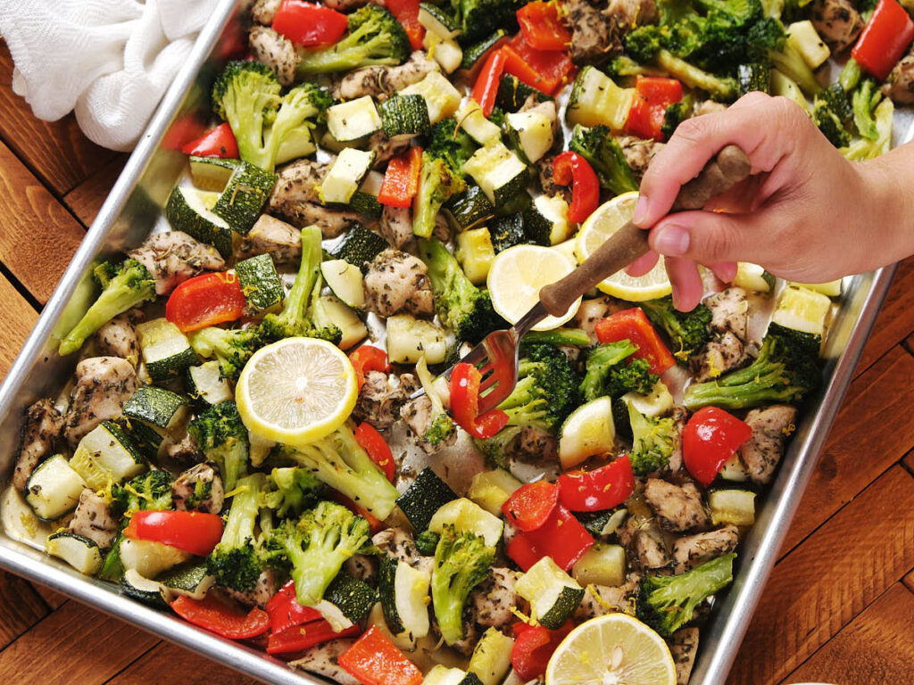 A hand uses a fork to pick up food from a baking tray filled with chicken and vegetables.