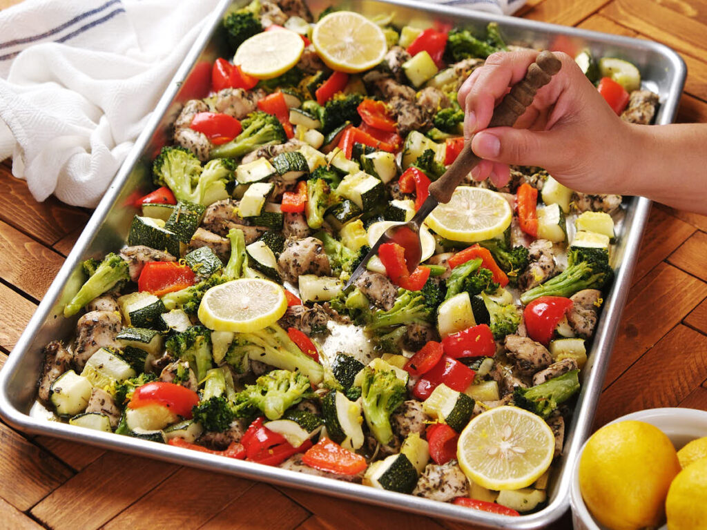 A hand stirs a sheet pan filled with roasted chicken, broccoli, zucchini, red bell peppers, and lemon slices.