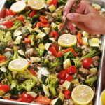 A hand holding a fork hovers over a baking sheet filled with roasted chicken, broccoli, zucchini, red bell peppers, and lemon slices.