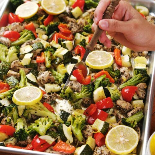 A hand holding a fork hovers over a baking sheet filled with roasted chicken, broccoli, zucchini, red bell peppers, and lemon slices.