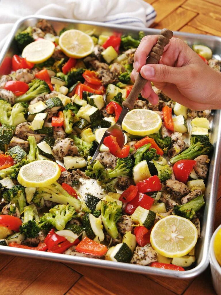 A hand holding a fork hovers over a baking sheet filled with roasted chicken, broccoli, zucchini, red bell peppers, and lemon slices.