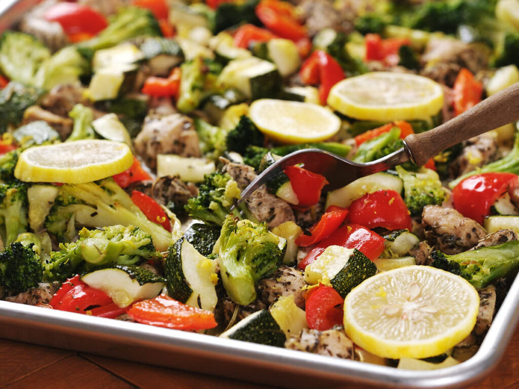 A sheet pan filled with roasted vegetables, including broccoli, zucchini, red bell peppers, and lemon slices, with a serving fork resting on top.