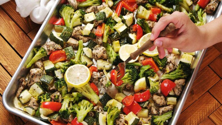 A hand uses a fork to serve a sheet pan meal with baked chicken, broccoli, zucchini, red bell pepper, and lemon slices on a metal tray.