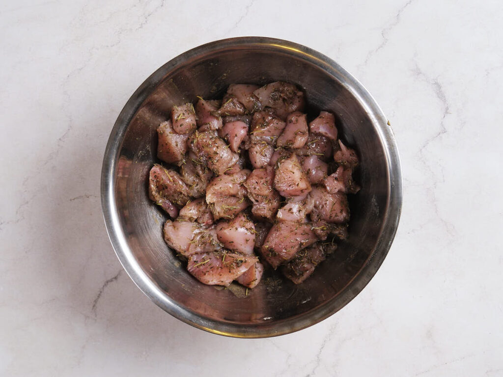 Raw, marinated chicken pieces in a stainless steel bowl on a light marble surface.