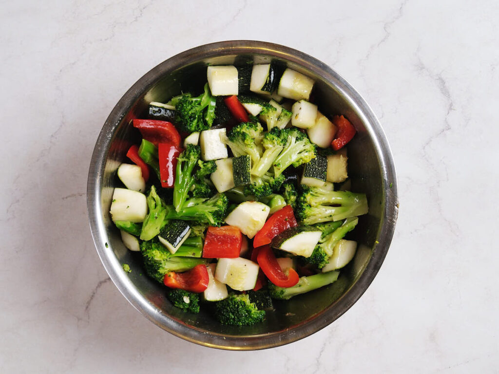 A metal bowl containing a mix of chopped broccoli, zucchini, and red bell pepper on a light marble surface.