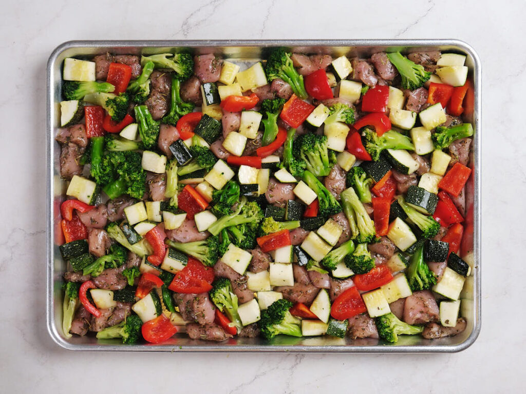 A sheet pan filled with diced chicken, broccoli, red bell pepper, and zucchini, all seasoned and ready for roasting.