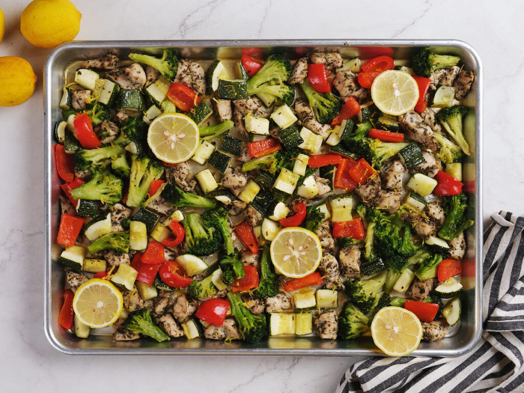 A sheet pan filled with chopped broccoli, zucchini, red bell pepper, pieces of chicken, and lemon slices.
