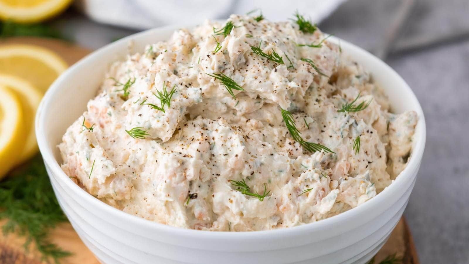 A white bowl filled with a creamy mixture, garnished with dill and ground pepper. Lemon slices are visible in the background.
