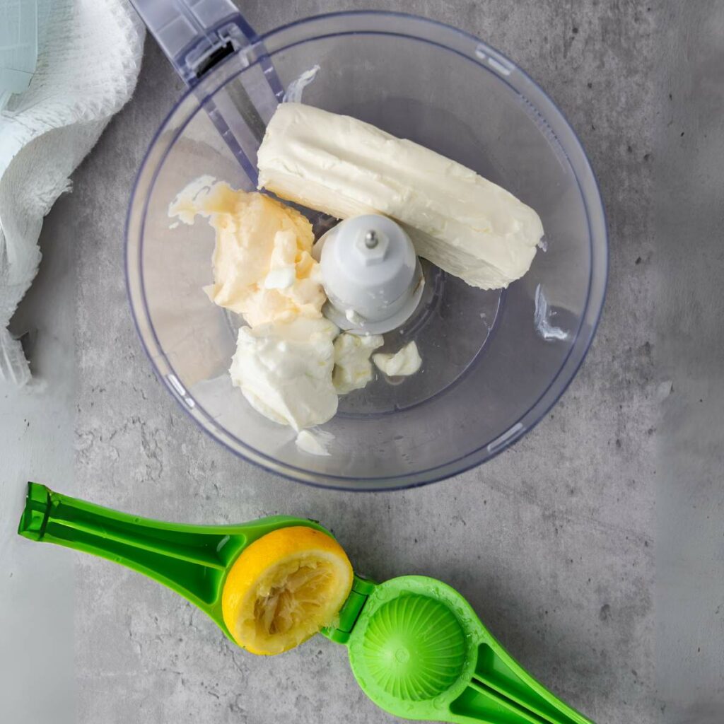 Food processor with cream cheese and mayo inside, next to a green citrus squeezer with a half-juiced lemon.