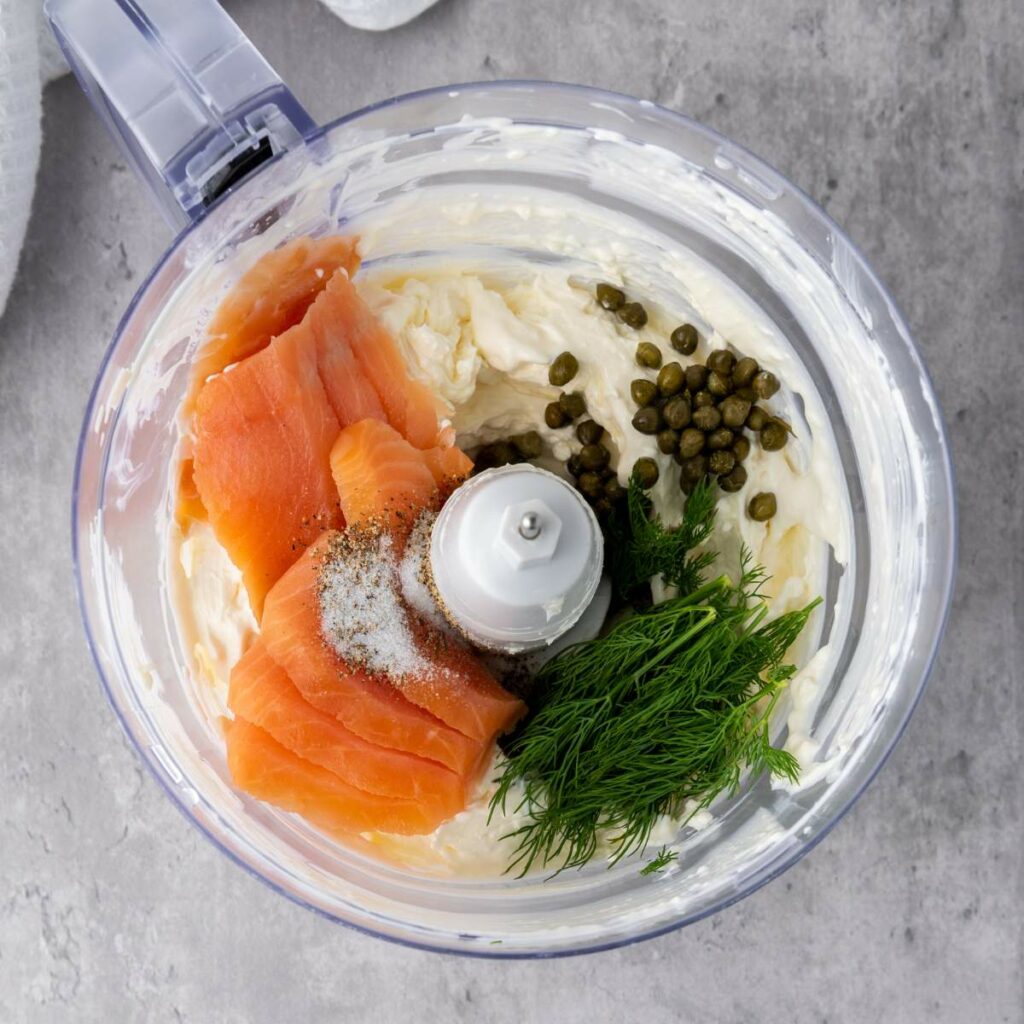Food processor bowl with cream cheese, smoked salmon, capers, fresh dill, and seasoning, ready to be blended.