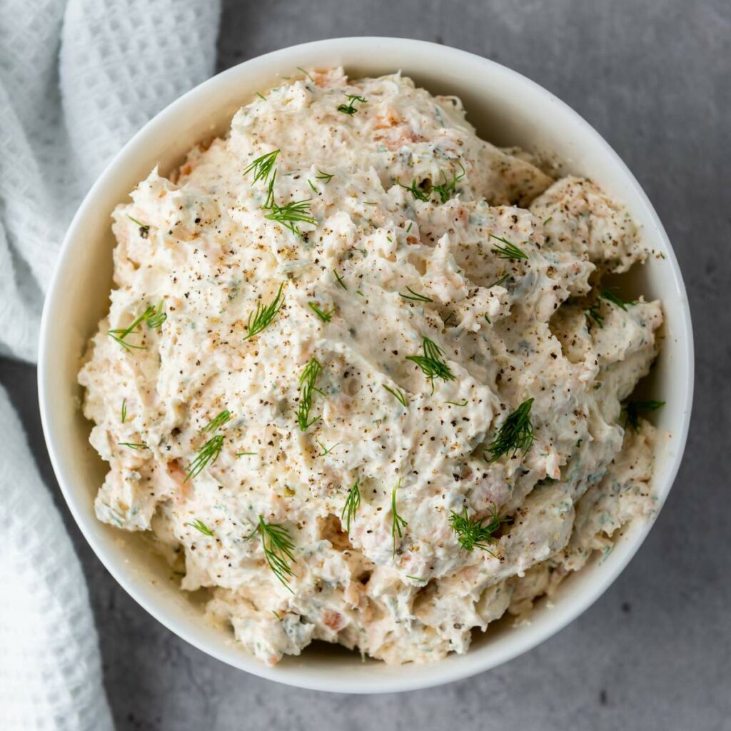 Bowl of creamy dip garnished with dill and black pepper, placed on a gray surface next to a white napkin.