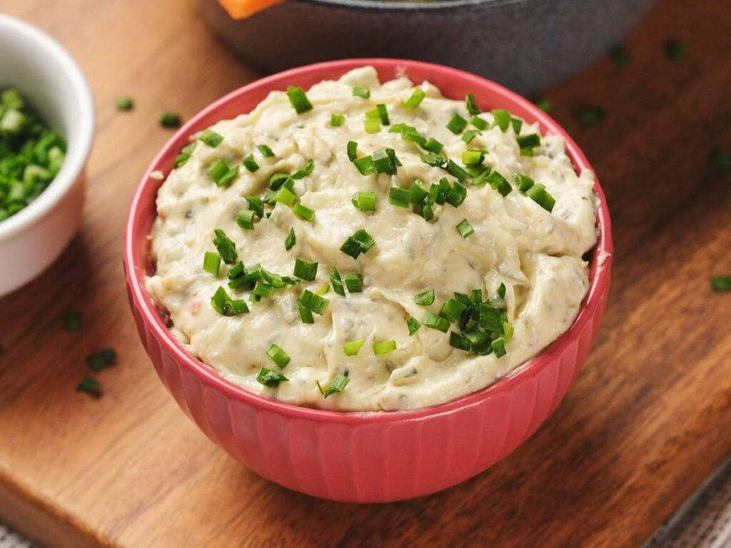 A pink bowl of sour cream dip topped with chopped green herbs on a wooden surface.