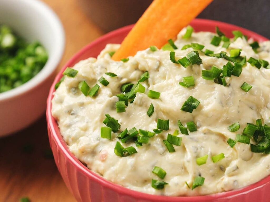 Bowl of sour cream dip garnished with chopped green onions, with a carrot stick dipped in.