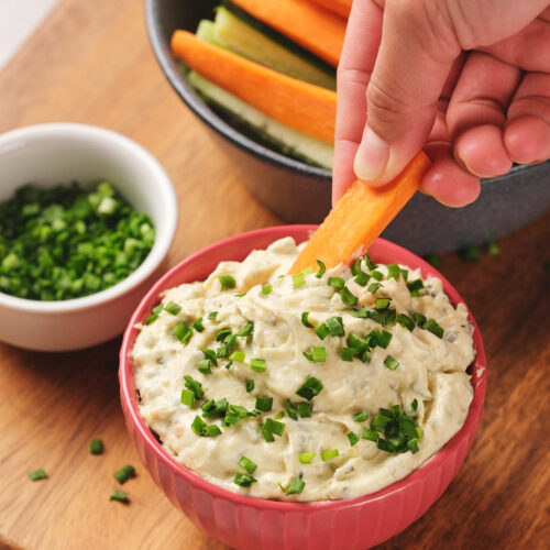 A hand dips a carrot stick into a bowl of sour cream dip topped with chopped chives.