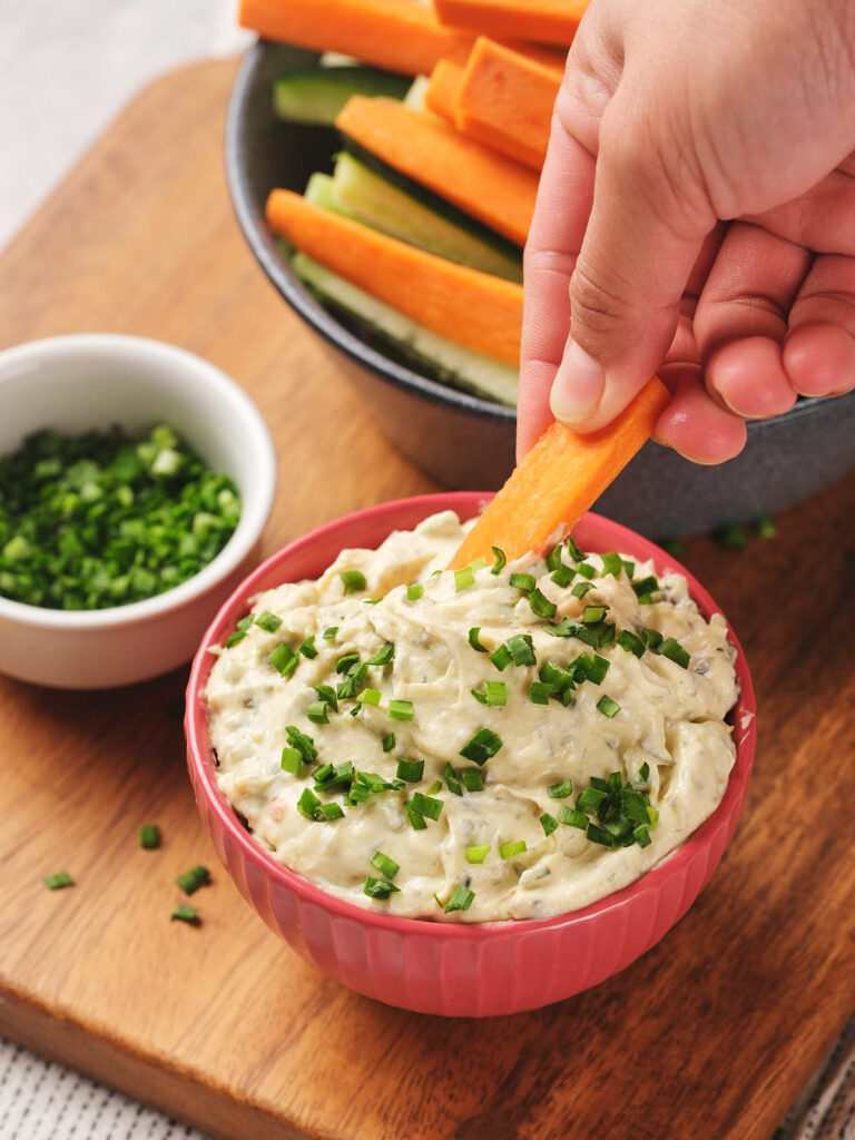 A hand dips a carrot stick into a bowl of sour cream dip topped with chopped chives.