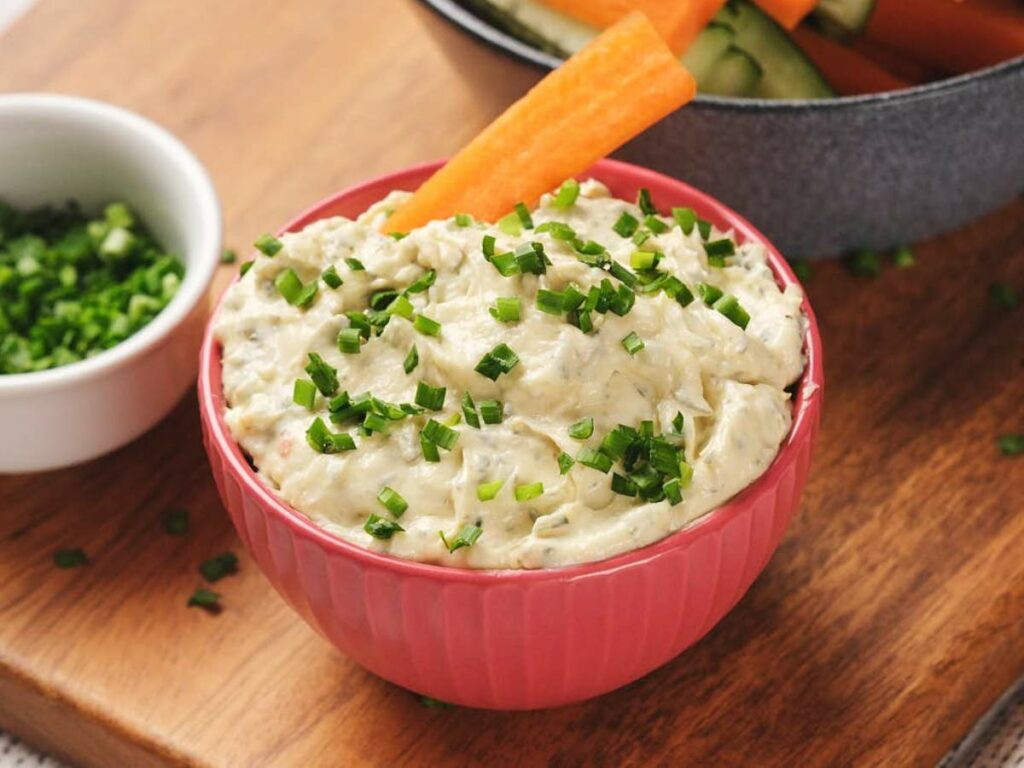 A pink bowl of sour cream dip topped with chopped chives, accompanied by carrot and cucumber sticks.
