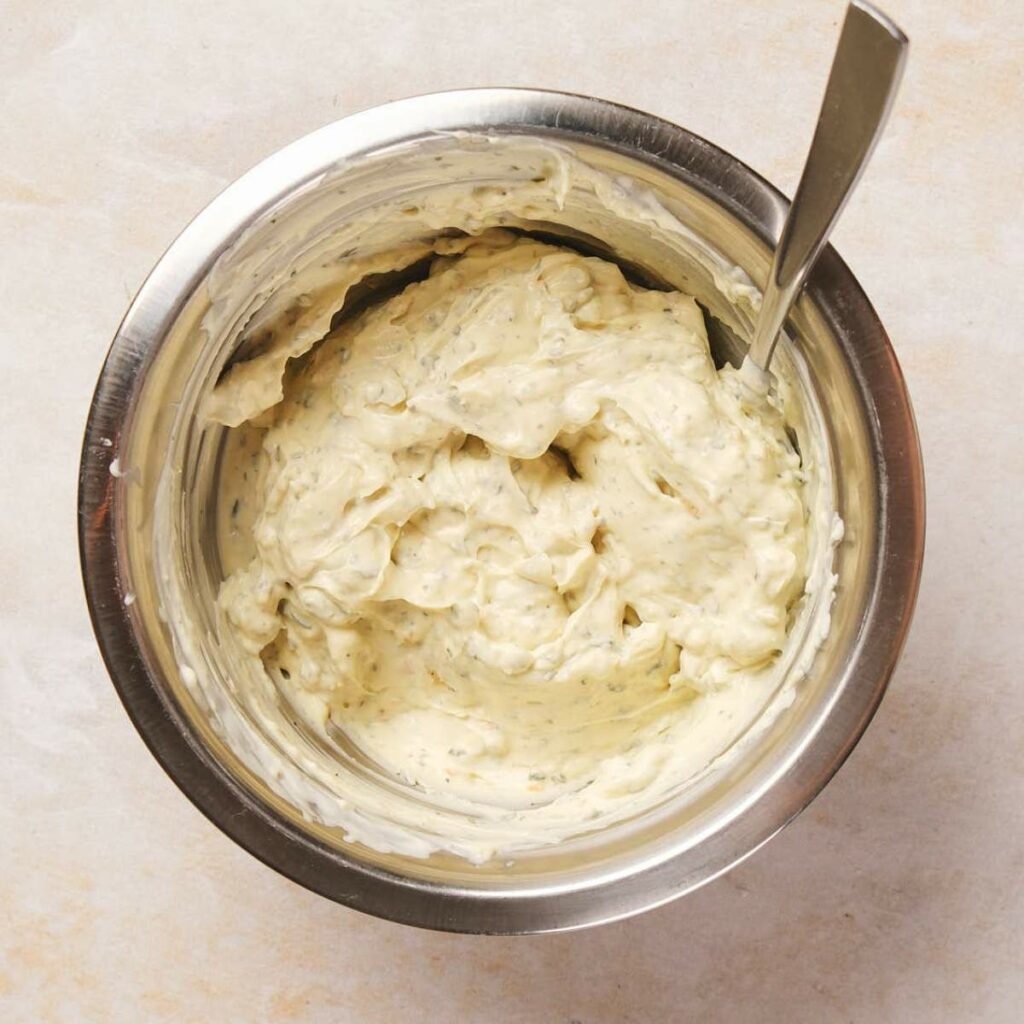 A stainless steel bowl filled with creamy, herb-infused dip. A metal spoon rests in the mixture.