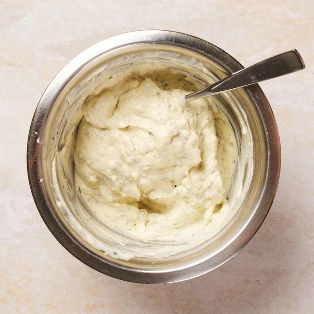 A stainless steel bowl filled with creamy white dressing, with a metal spoon resting inside.