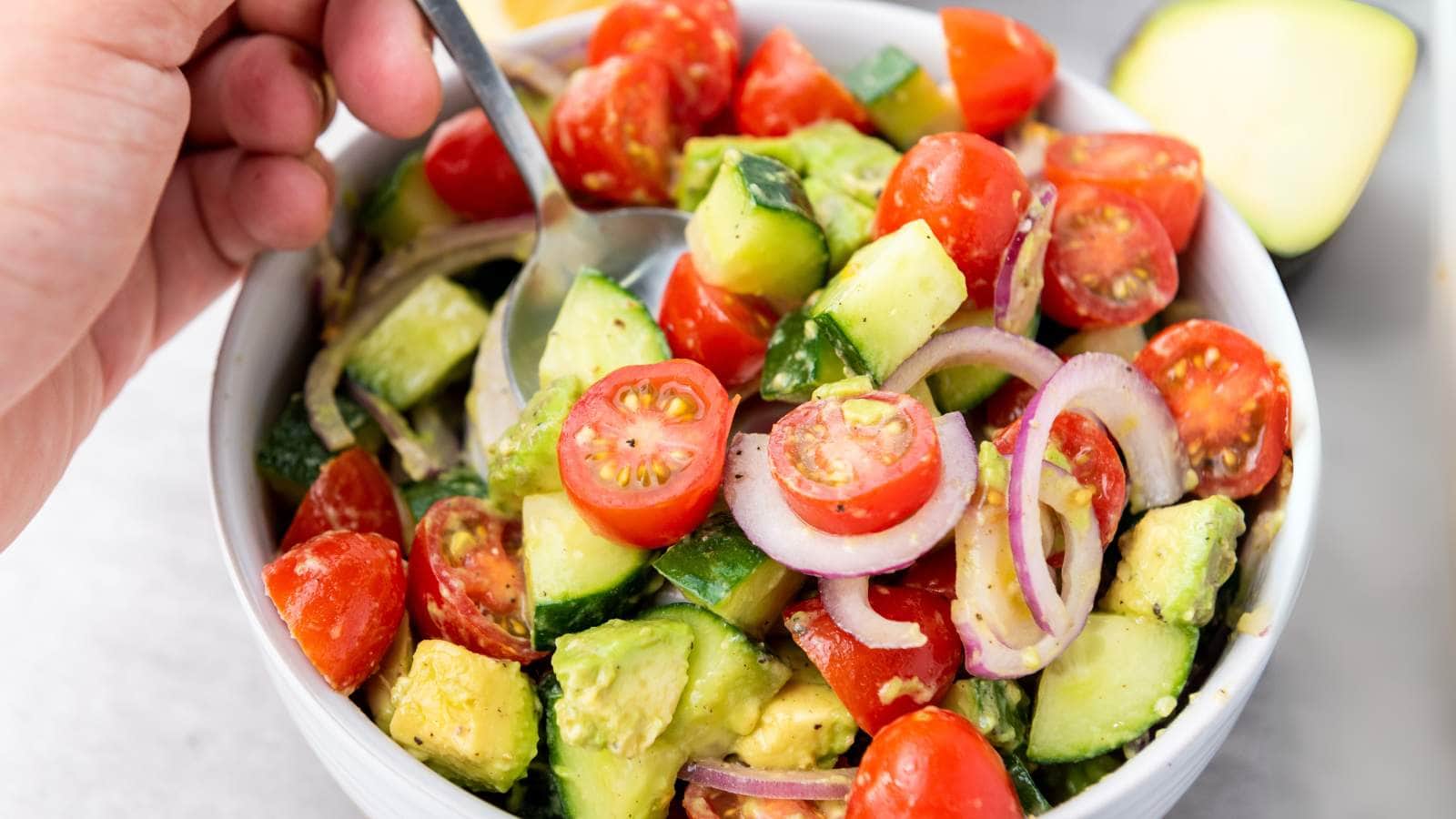 A hand holds a spoon in a bowl of avocado tomato salad with cucumber.