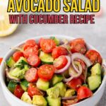 A bowl of avocado salad with cherry tomatoes, cucumber, and red onion slices on a light surface.