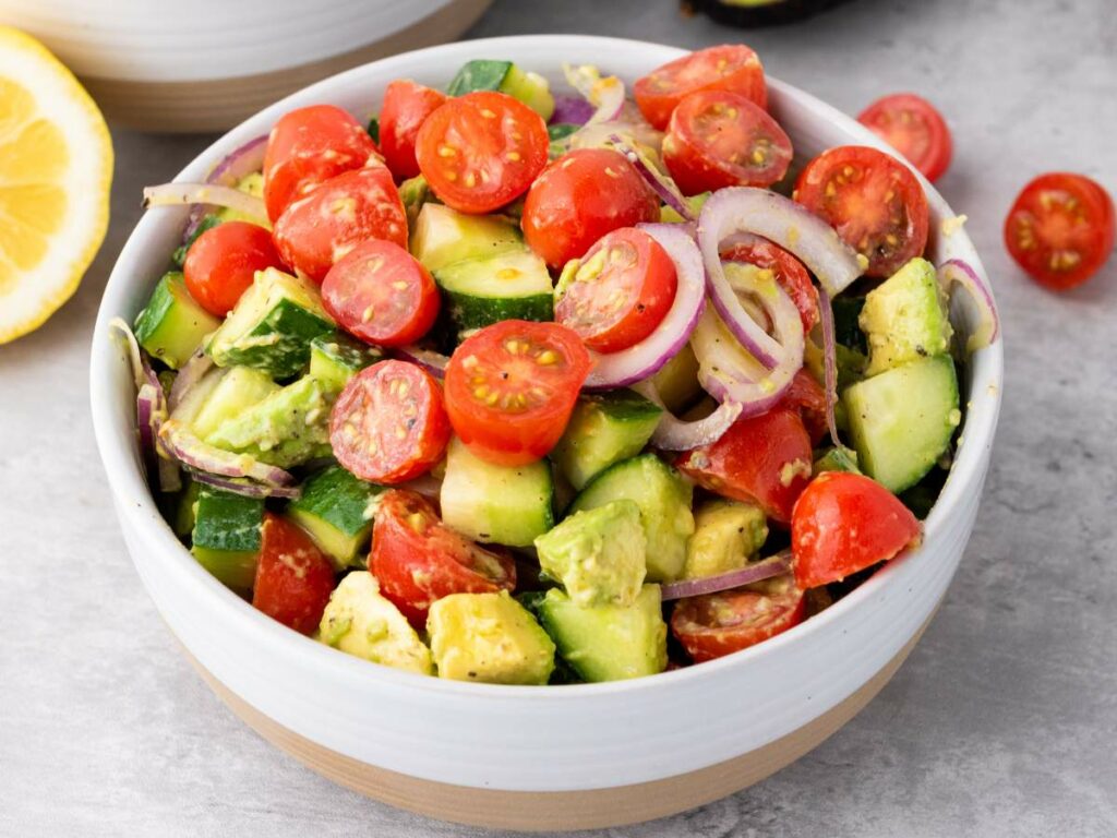 A bowl of avocado tomato salad with cucumbers on a light surface.