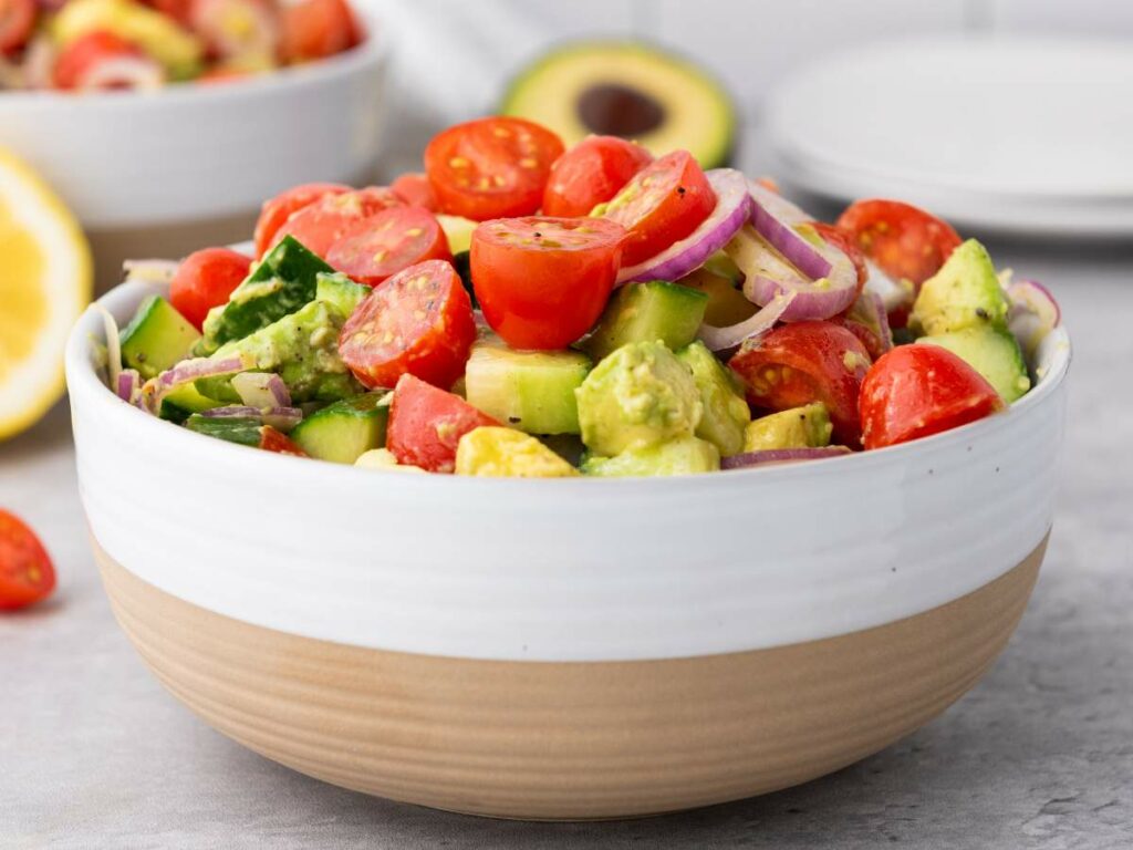 A bowl of salad containing cherry tomatoes, cucumber, avocado, and red onion slices on a light surface.
