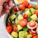 A hand holding a spoon over a bowl of cucumber, tomato, and avocado salad.