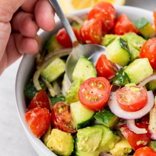 A hand holding a spoon over a bowl of cucumber, tomato, and avocado salad.