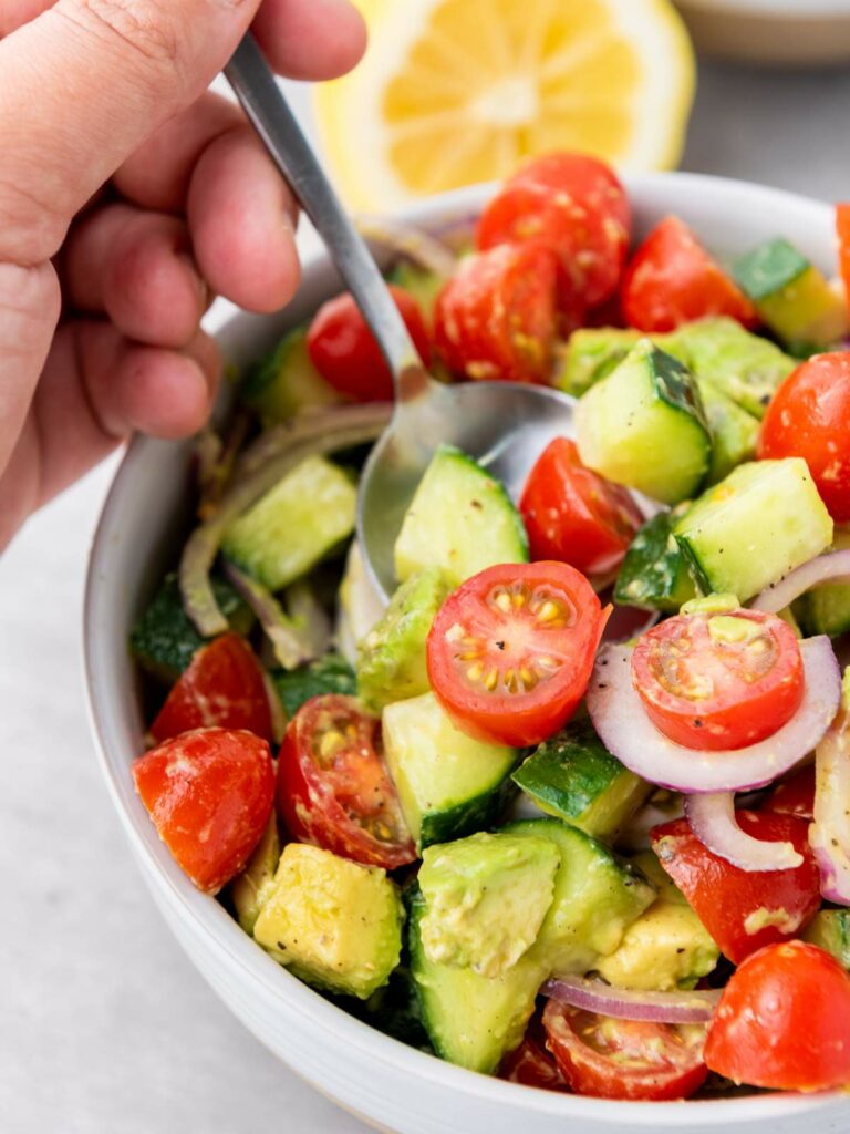A hand holding a spoon over a bowl of cucumber, tomato, and avocado salad.
