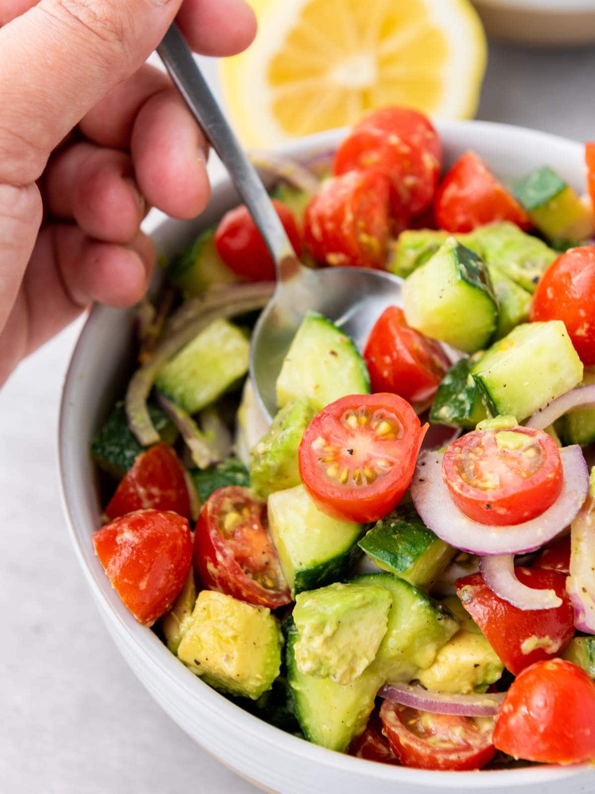 A hand holding a spoon over a bowl of cucumber, tomato, and avocado salad.