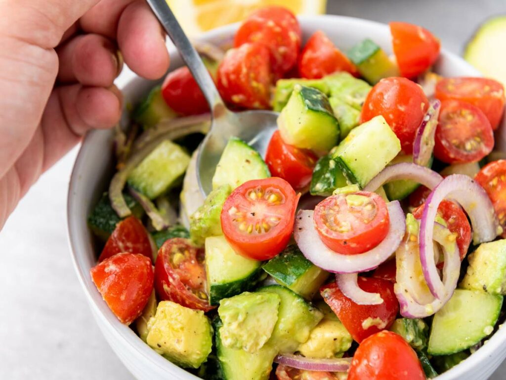 A hand holds a spoon in a bowl of salad with cherry tomatoes, cucumber, avocado, and red onion slices.