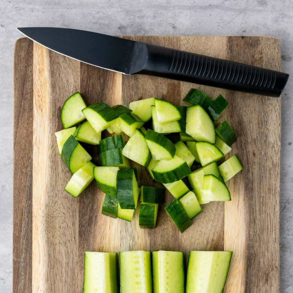 A black-handled knife and chopped cucumber pieces are on a wooden cutting board.