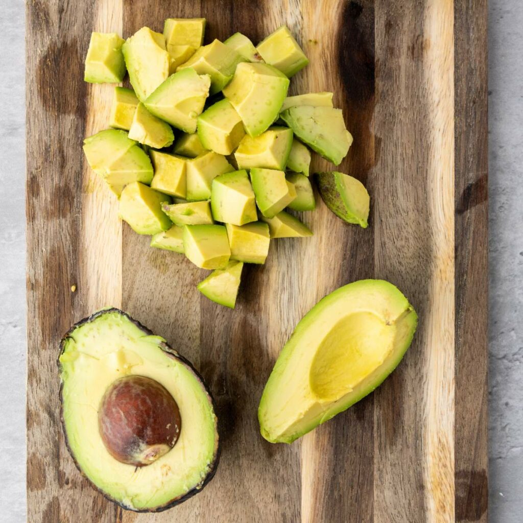 A halved avocado with seed, a halved avocado without seed, and diced avocado pieces on a wooden cutting board.