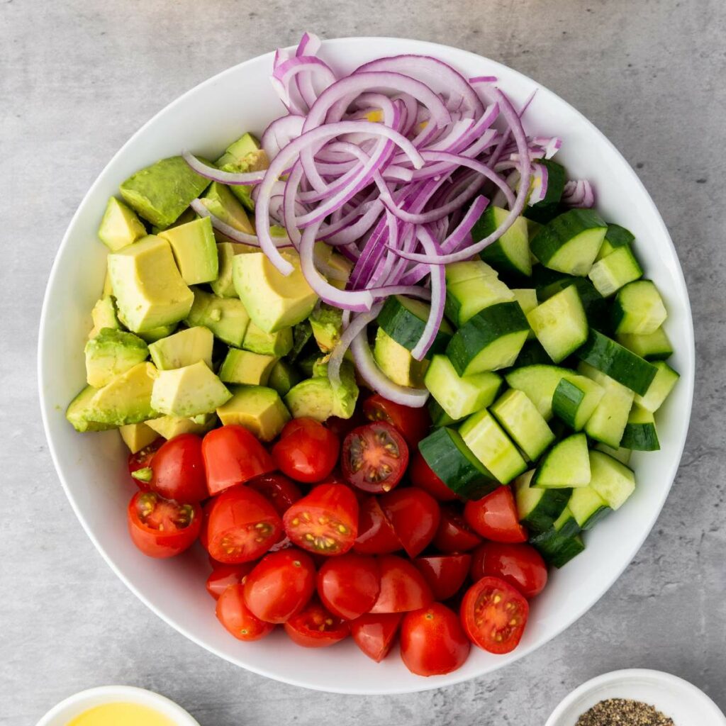 A bowl containing chopped avocado, sliced red onion, cubed cucumber, and halved cherry tomatoes on a gray surface.