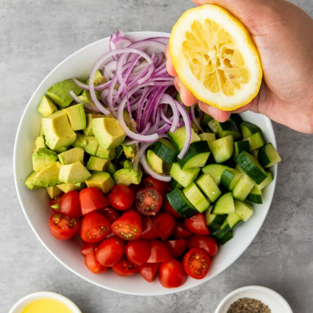 A hand squeezes half a lemon over a bowl filled with chopped avocado, cucumber, cherry tomatoes, and red onion.