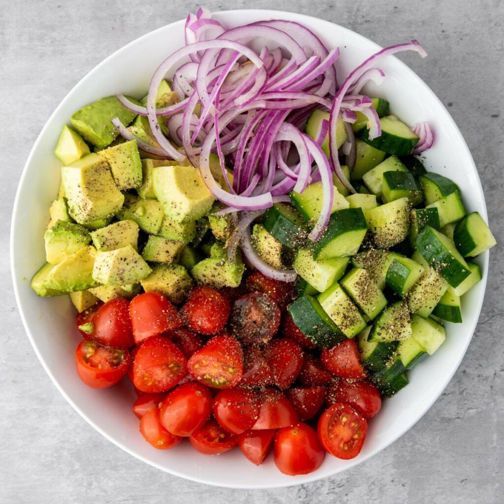 A white bowl filled with chopped avocado, cherry tomatoes, cucumber, and sliced red onion, topped with ground black pepper.