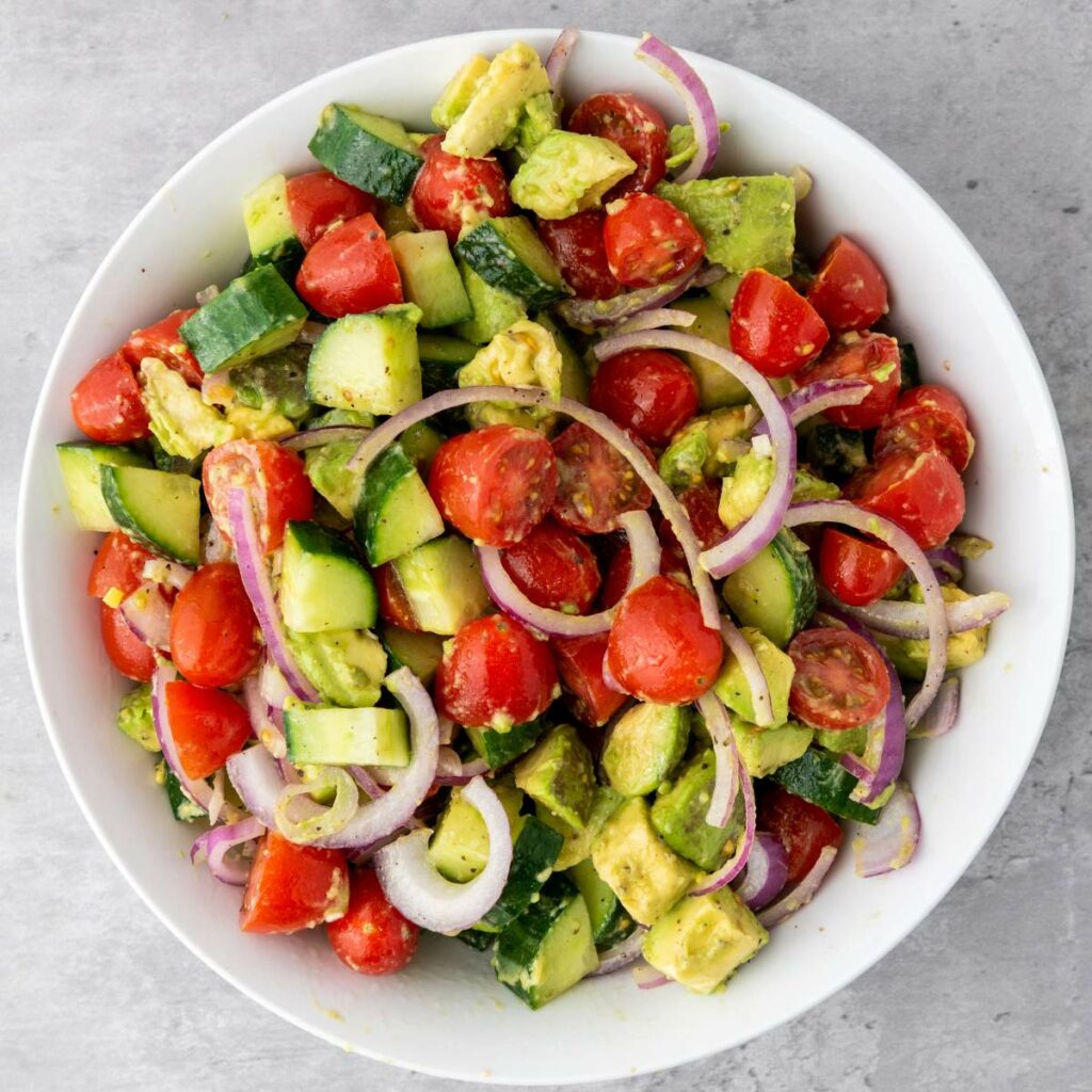 A white bowl filled with a salad of chopped cucumber, cherry tomatoes, avocado, and sliced red onion on a gray surface.