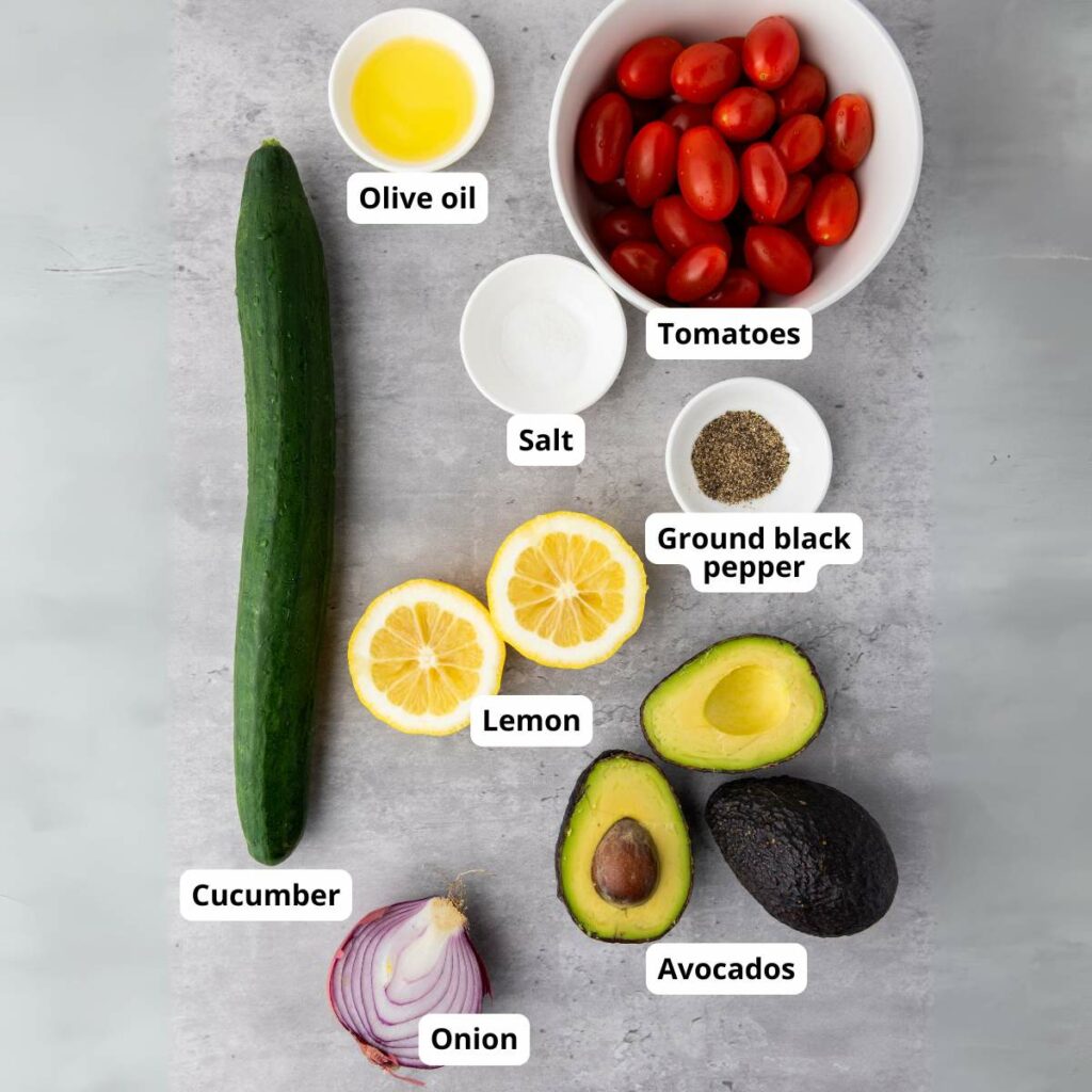 Ingredients for avocado tomato salad with cucumber arranged on a gray surface, each labeled.