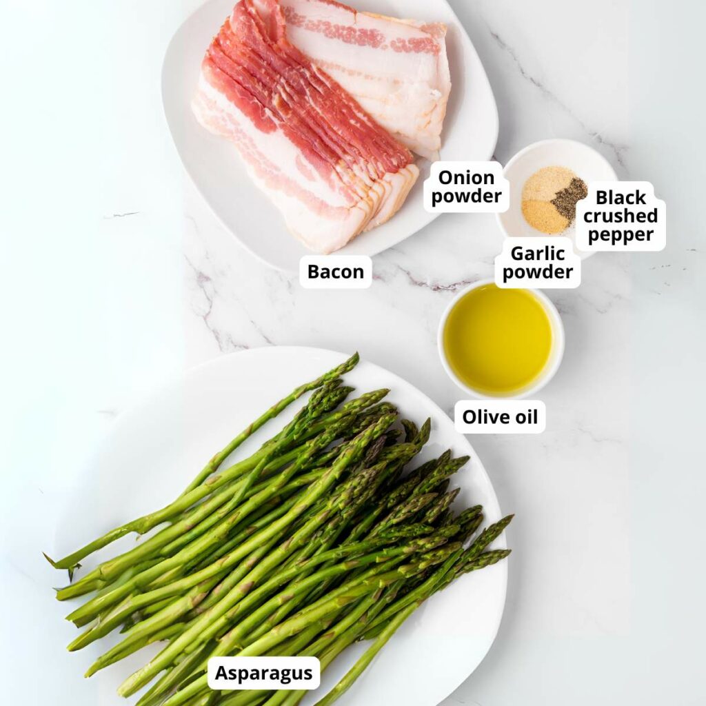 Raw asparagus, uncooked bacon slices, and small bowls of onion powder, black crushed pepper, garlic powder, and olive oil on white plates on a marble surface.
