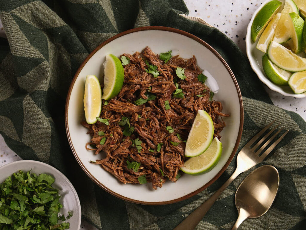 A plate of beef barbacoa garnished with lime wedges and chopped cilantro.