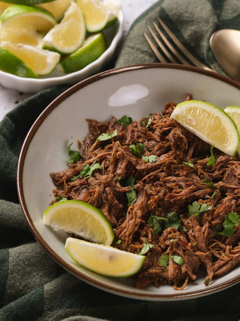 A bowl of beef barbacoa garnished with lime wedges and chopped herbs.