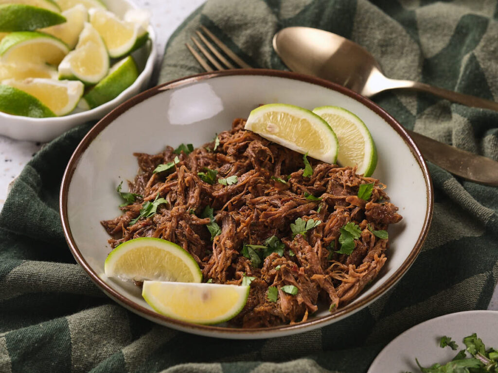 A bowl of beef barbacoa garnished with lime wedges and chopped cilantro.