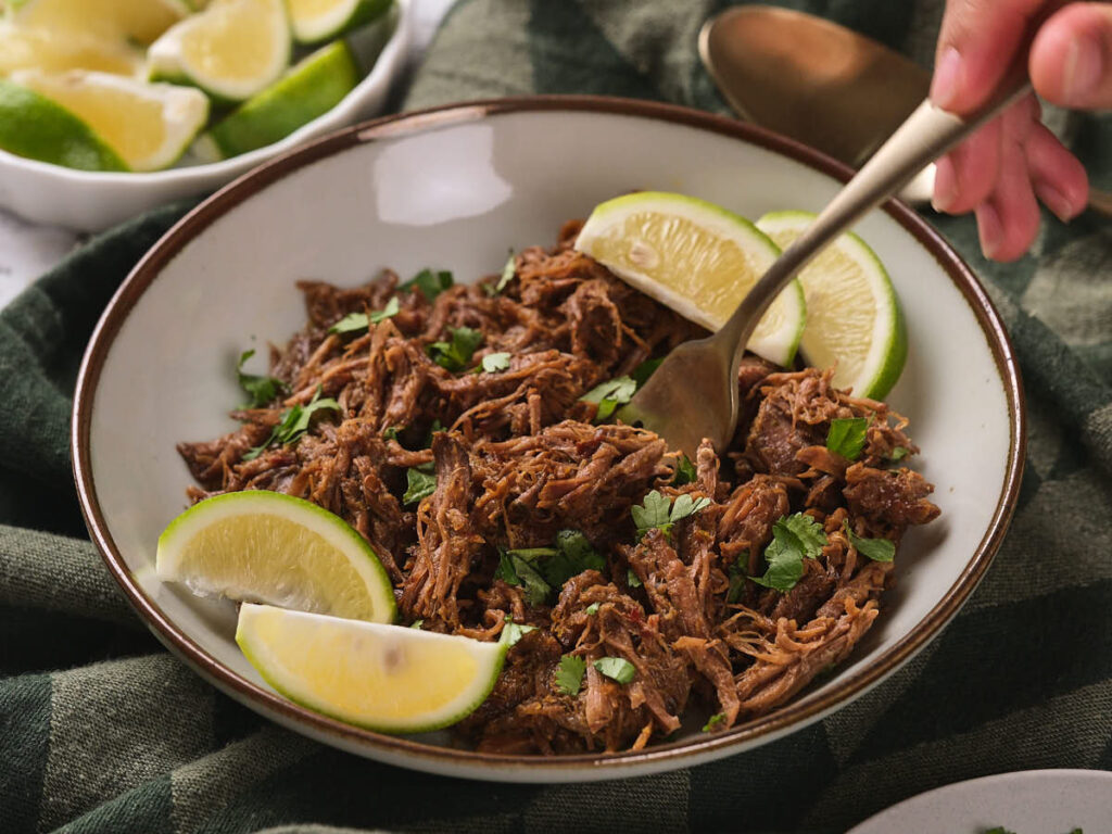 A bowl of beef barbacoa garnished with chopped cilantro and lime wedges.