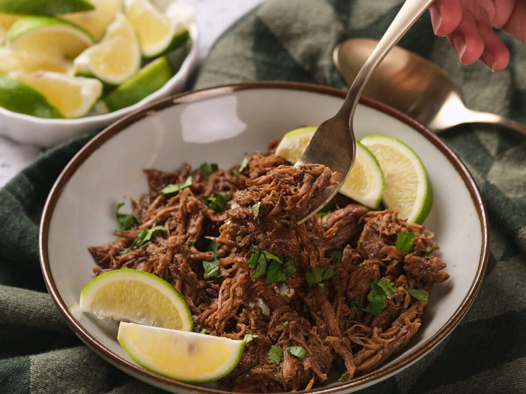 A bowl of beef barbacoa garnished with lime wedges and chopped herbs, with a hand holding a spoon.