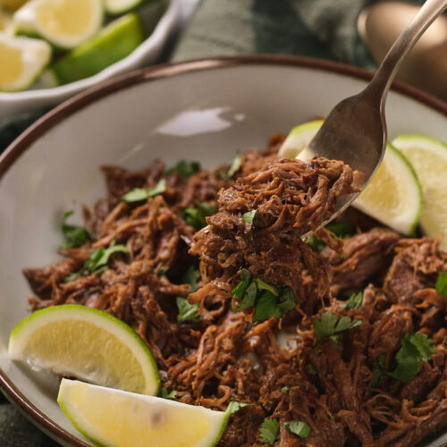 A bowl of beef barbacoa garnished with chopped cilantro and lime wedges.